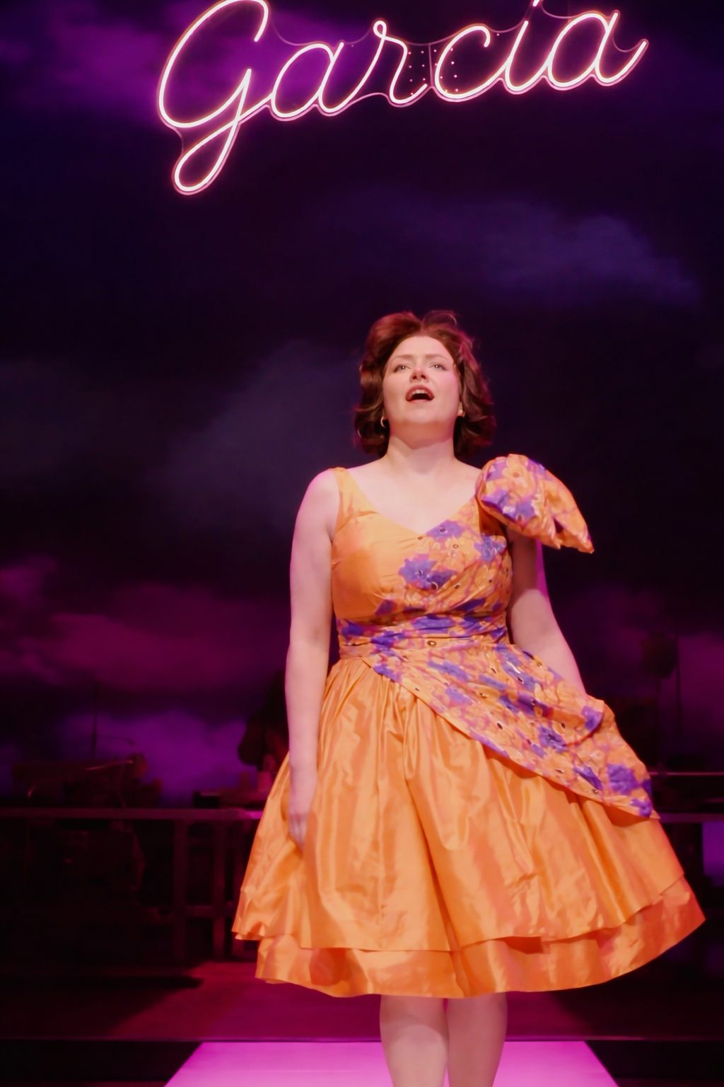 Gloria Vivica Benavides on stage in an orange dress beneath a Garcia neon sign
